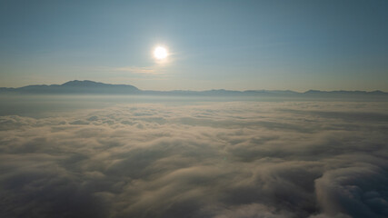 A serene view of the early morning sun rising above a vast sea of clouds, with distant mountain peaks faintly visible on the horizon, creating a peaceful and ethereal landscape. Flying over the cloud