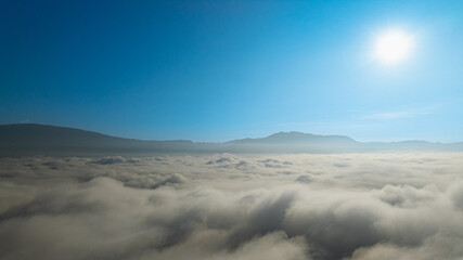 A serene view of the early morning sun rising above a vast sea of clouds, with distant mountain peaks faintly visible on the horizon, creating a peaceful and ethereal landscape. Flying over the cloud