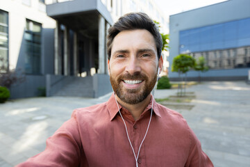 A man smiles during a video call, holding his phone. He wears earphones while standing in an urban...
