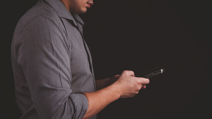 A man in gray shirt in dark black background is holding a mobile phone in his hand and typing message email on smartphone in concept of communication technology.
