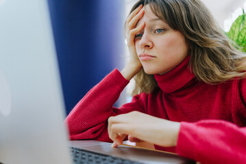 Young woman feeling tired and exhausted while working remotely from her home office, struggling to...