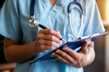 Close-up of hands holding clipboard, pen writing medical notes,  blue scrubs and stethoscope visible, representing healthcare and medical professionalism