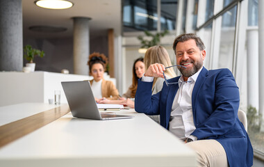 Businessman holding eyeglasses and smiling at camera while female coworkers working in the background in boardroom