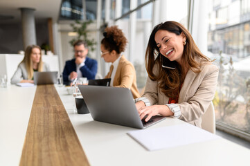 Smiling lawyer talking over mobile phone and typing e-mail on laptop in board room with coworkers...