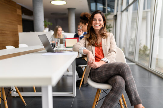 Portrait of confident businesswoman with laptop at table sitting in board room while colleagues collaborating in the background