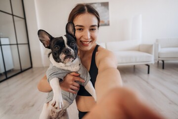 Doing selfie. Sportive woman in yoga clothes is with pug dog at home