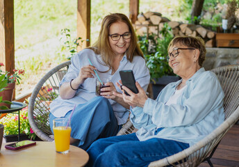 Two mature women friends mother and daughter using smartphone while sitting on sunny terrace of...
