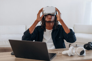User is with headset. Young man is sitting by table with laptop and virtual reality glasses