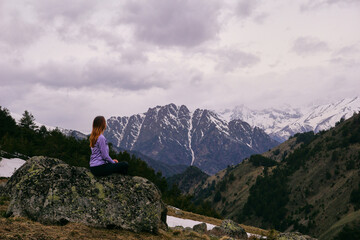 Naklejka premium A young woman sits on a large rock and looks at the mountains. Active travel in nature, hiking in the mountains