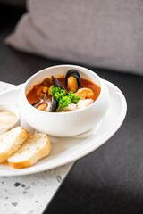 A bowl of seafood soup with mussels and shrimp in a rich tomato broth, garnished with parsley, served with bread and sauce on a white plate.