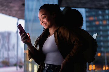 Stylish young female Latina checks exclusive online discounts on her cellphone while standing near glass building. reflection adds depth, emphasizing modern digital connectivity. Technology using
