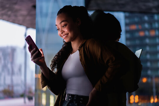 Stylish young female Latina checks exclusive online discounts on her cellphone while standing near glass building. reflection adds depth, emphasizing modern digital connectivity. Technology using