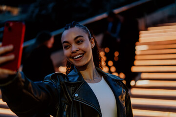 Smiling attrractive Hispanic woman in black leather jacket take selfie for social media or enjoying video call in vibrant city at night.Warm urban lights create stylish and modern social media moment