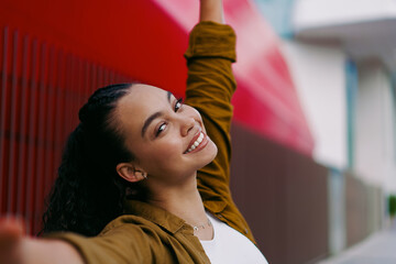Cheerful young Latina woman raises arms with bright smile in urban setting.Female joyful expression represents happiness, confidence, and positivity.Concept of self-love, freedom, and modern lifestyle