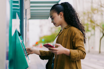 Focused Latina woman using self-service kiosk at transit station while holding a smartphone. Female tourist represents modern transportation, digital solutions, and convenience in everyday city life