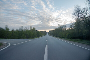 Fototapeta premium Empty countryside road stretching into the distance under a cloudy blue sky, flanked by green trees at dawn or dusk.