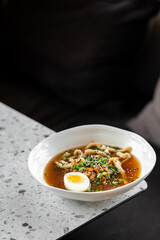 A bowl of ramen soup with noodles, vegetables, and a halved boiled egg, garnished with chopped green onions and sesame seeds, placed on a speckled countertop.