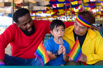 african and asian male gay couple sitting on beach chair are taking care boy kid son eating ice cream while travel on summer trip