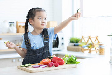 cute asian little child girl eating slice a cucumber with different vegetables in the kitchen