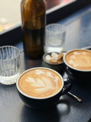 A warm cup of latte with delicate foam art on a dark table, surrounded by glassware and sugar cubes. Soft morning light highlights a calm and cozy breakfast atmosphere.
