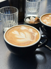 A warm cup of latte with delicate foam art on a dark table, surrounded by glassware and sugar cubes. Soft morning light highlights a calm and cozy breakfast atmosphere.
