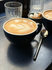 A warm cup of latte with delicate foam art on a dark table, surrounded by glassware and sugar cubes. Soft morning light highlights a calm and cozy breakfast atmosphere.