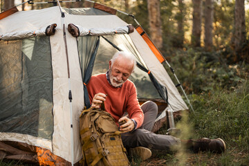 Senior Man Camping in Forest and Organizing His Backpack Outside Tent