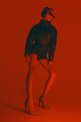 Low angle full-length portrait of stylish young woman in sunglasses, jacket, short and heels, posing confidently against red studio background in neon light. Concept of fashion, style, youth culture