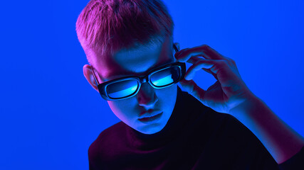 Portrait of stylish young boy in sunglasses and black turtleneck posing against blue background in neon light. Close-up. Concept of bold fashion, style, youth culture, self-expression