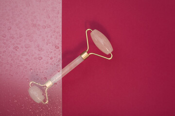 Pink jade roller for aesthetic facial massage on burgundy background with water drops. Self-care...