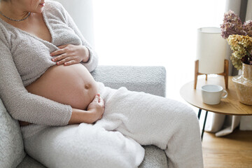 Pregnant woman hugging baby bump, sitting and relaxing on cozy couch in modern living room. Happy Mothers day. Love and care concept. Fertility and maternity