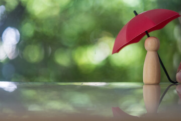 One wooden figure stands protected under a red umbrella by human hand against a natural background, symbolizing safety and security. insurance and social protection patronage and mentoring