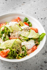 Fresh salad with cucumber, tomato, mixed greens, sesame seeds, sunflower seeds, and creamy dressing in a white bowl on a speckled gray surface