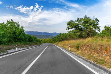 A winding asphalt road curves gently through a green countryside landscape under a vibrant blue sky...