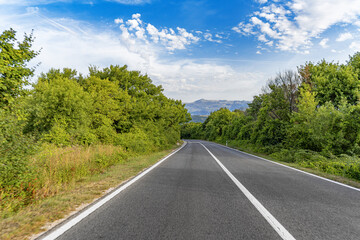 A winding asphalt road curves gently through a green countryside landscape under a vibrant blue sky with scattered clouds and distant mountains.