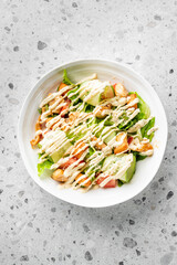 Fresh vegetable salad with lettuce, cucumbers, tomatoes, croutons, and cashews, topped with creamy dressing and sesame seeds, served in a white bowl on a terrazzo countertop.