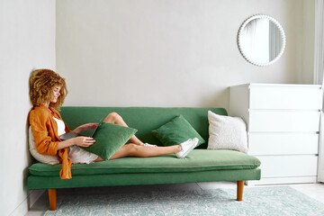 Young woman relaxing on a green couch in a cozy living room, wearing a casual outfit with a...