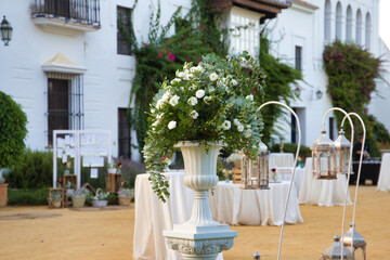 Flower pot with eucalyptus branches and white flowers in the decoration for an outdoor wedding. There are catering tables and lanterns in the decoration of the event.