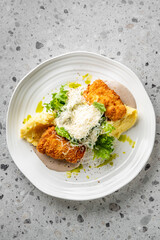 A gourmet dish featuring crispy breaded chicken, mashed potatoes, fresh greens, and grated cheese, garnished with a drizzle of green sauce, presented on a white plate with a textured grey background.