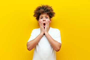 shocked curly african american boy looking at camera in amazement over yellow isolated background,...