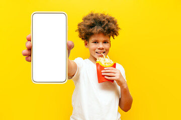 Cheerful curly african american man eating french fries and showing blank smartphone screen on yellow isolated background, hungry teenager holding fried potatoes and advertising fast food on internet