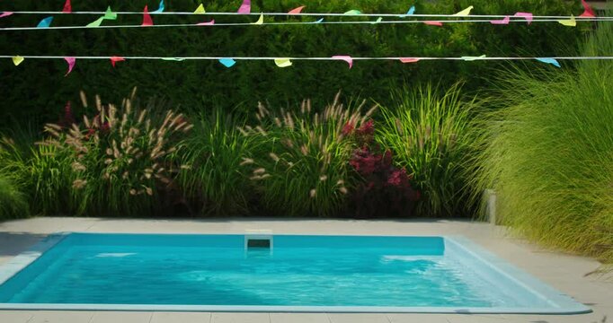 Empty backyard pool with colorful party flags above, surrounded by tall green and ornamental grass in a summer garden