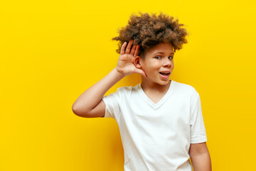 curious curly african american boy eavesdropping and holding hand near ear on yellow isolated...