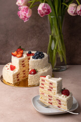 Strawberry and coconut birthday cake with cream and fresh berries. A slice of homemade layered birthday cake. In the background is a bouquet of pink tulips. Selective focus, close-up.