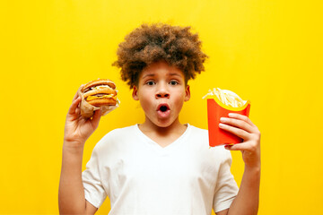surprised african american boy holding and showing fast food on yellow isolated background, shocked curly teenager advertising burger and fries in amazement