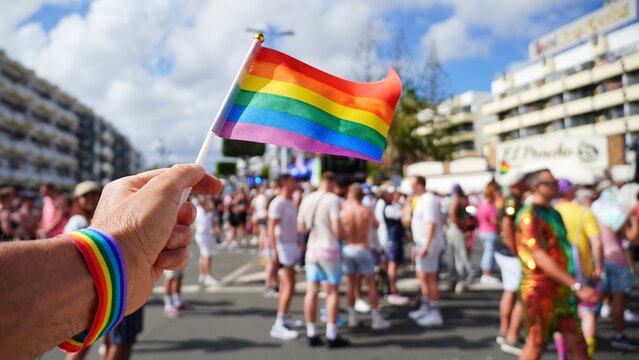 Hand holding rainbow flag and wearing rainbow wristband waves at Maspalomas Gay Pride parade celebrating pride month in Gran Canaria, Spain, with blurred crowd of tourists and locals in background