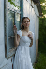 Young woman in a white dress standing by a rustic window, exuding a serene and dreamy emotion in a...