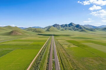 Aerial view of railway tracks traversing a vast, verdant landscape under a clear blue sky, with rolling hills in the background