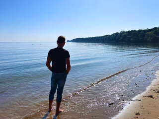 Slim man in jeans and top and bare feet on sandy Studland beach looking out to Old Harry Rock in...