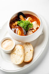 A gourmet seafood soup with mussels, shrimp, and fish, garnished with fresh parsley, served with crusty bread and creamy sauce on a white plate.